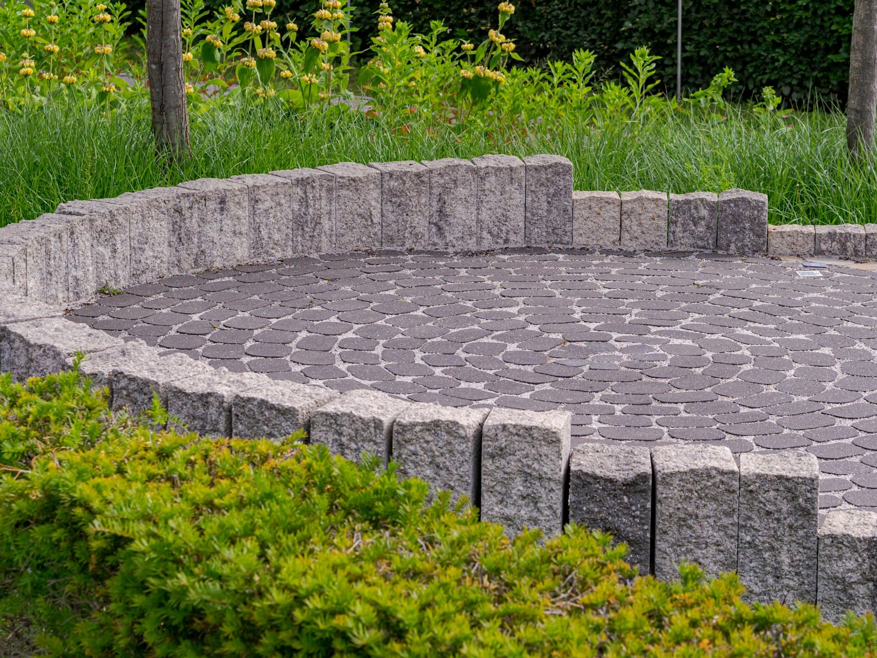 Circular paver patio in a fan pattern, edged with a low stone palisade wall and surrounded by green plantings