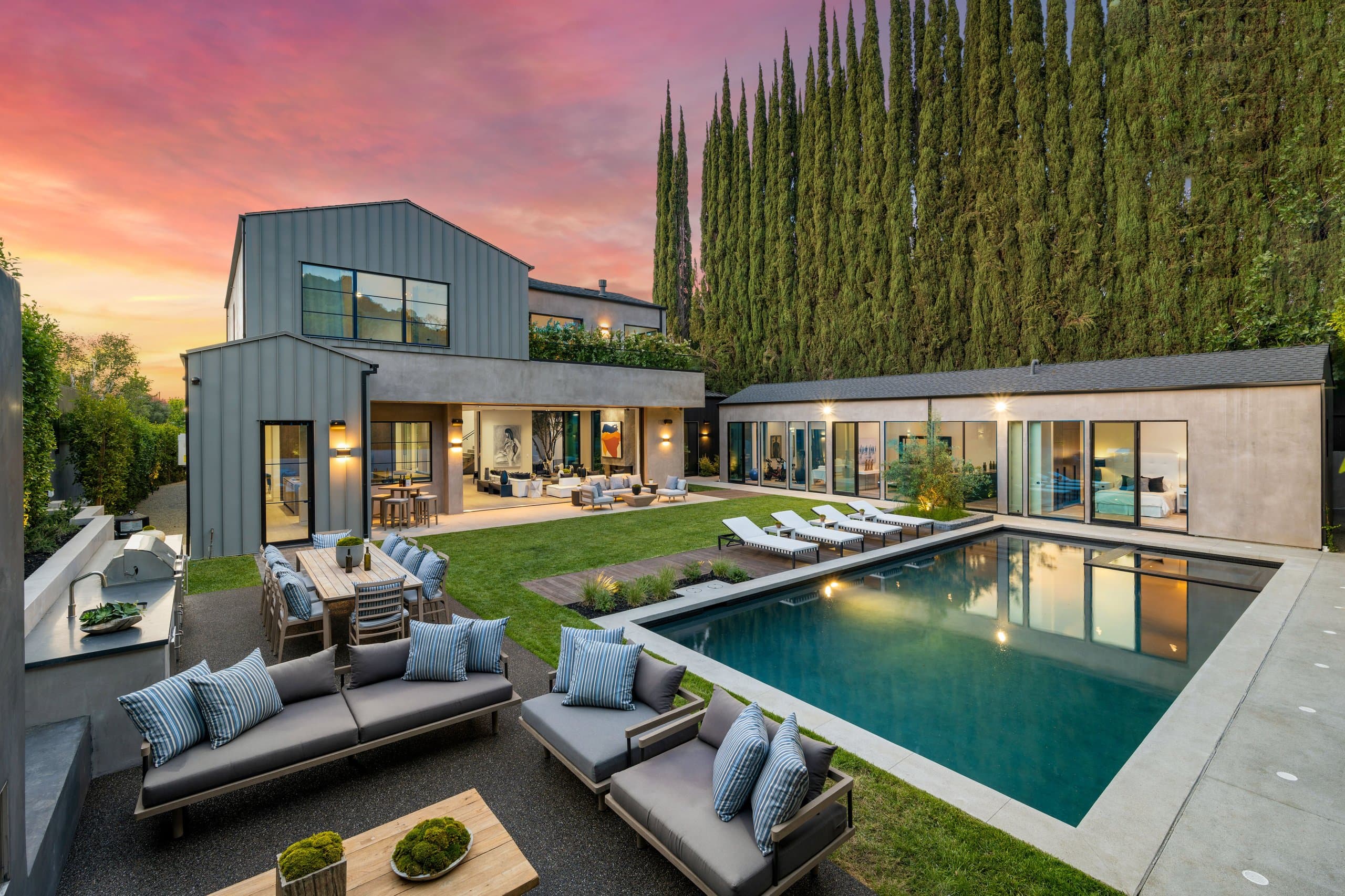 Modern dark home with backyard pool, lawn, loungers, and outdoor dining at dusk