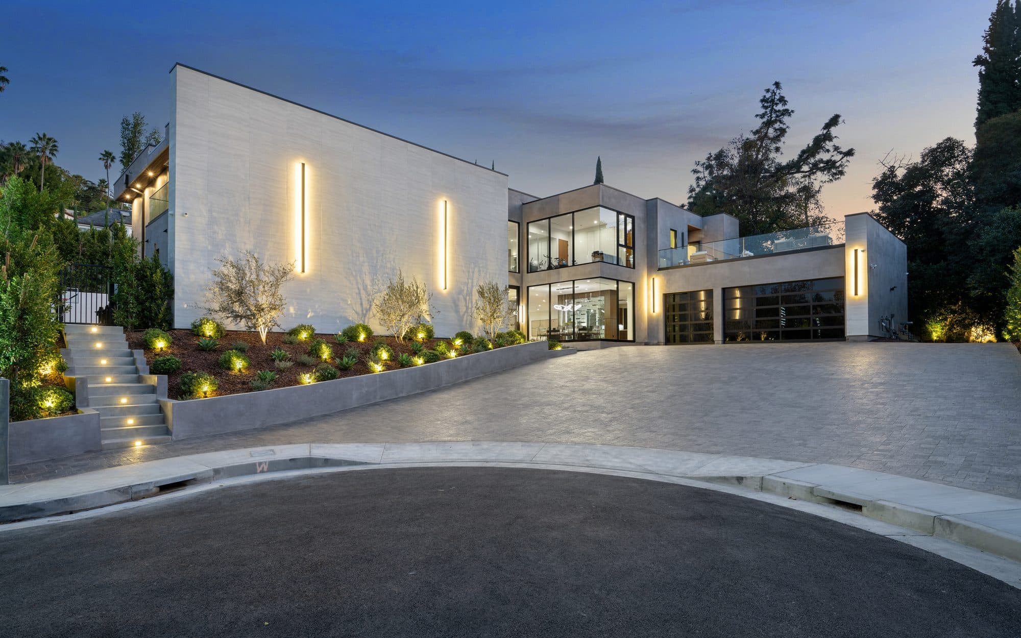 Curved estate paver driveway at dusk with terraced planters and architectural lighting