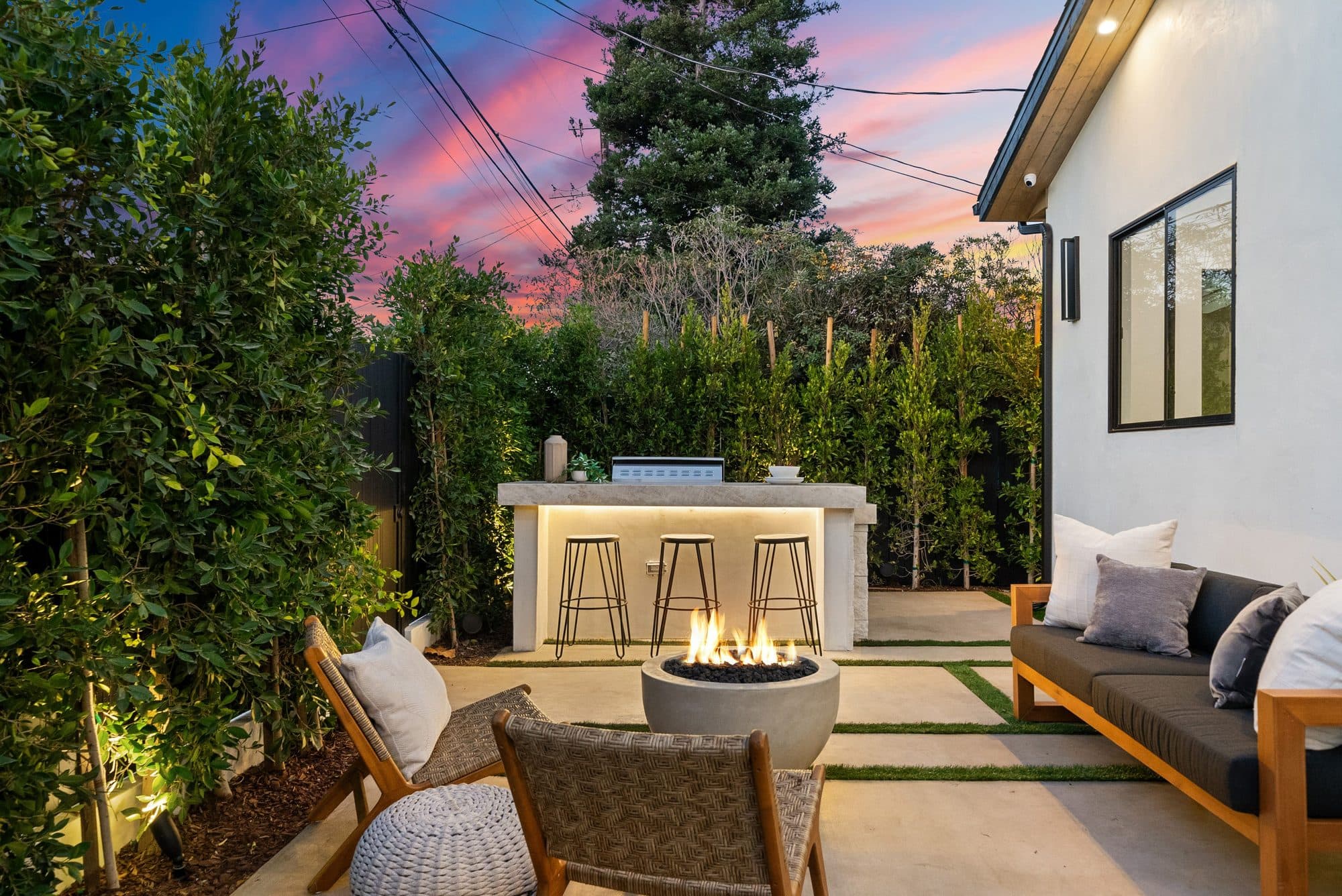 Backyard courtyard at sunset with a concrete fire bowl, bar seating, and lit privacy hedges