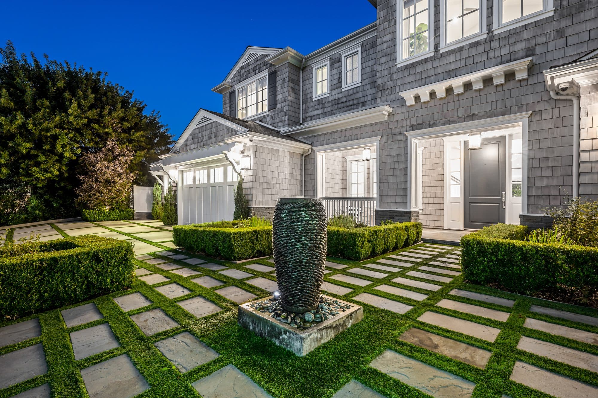 Gray shingle home facade with turf-grid pavers, boxwood hedges, and a stone water feature