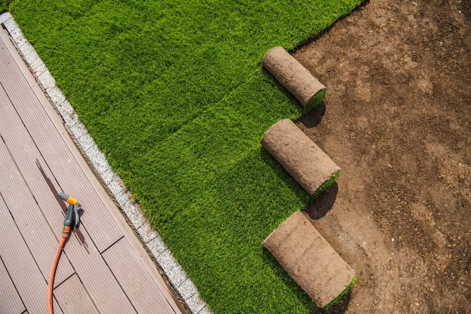 Aerial view of sod rolls being unrolled across graded soil beside a deck