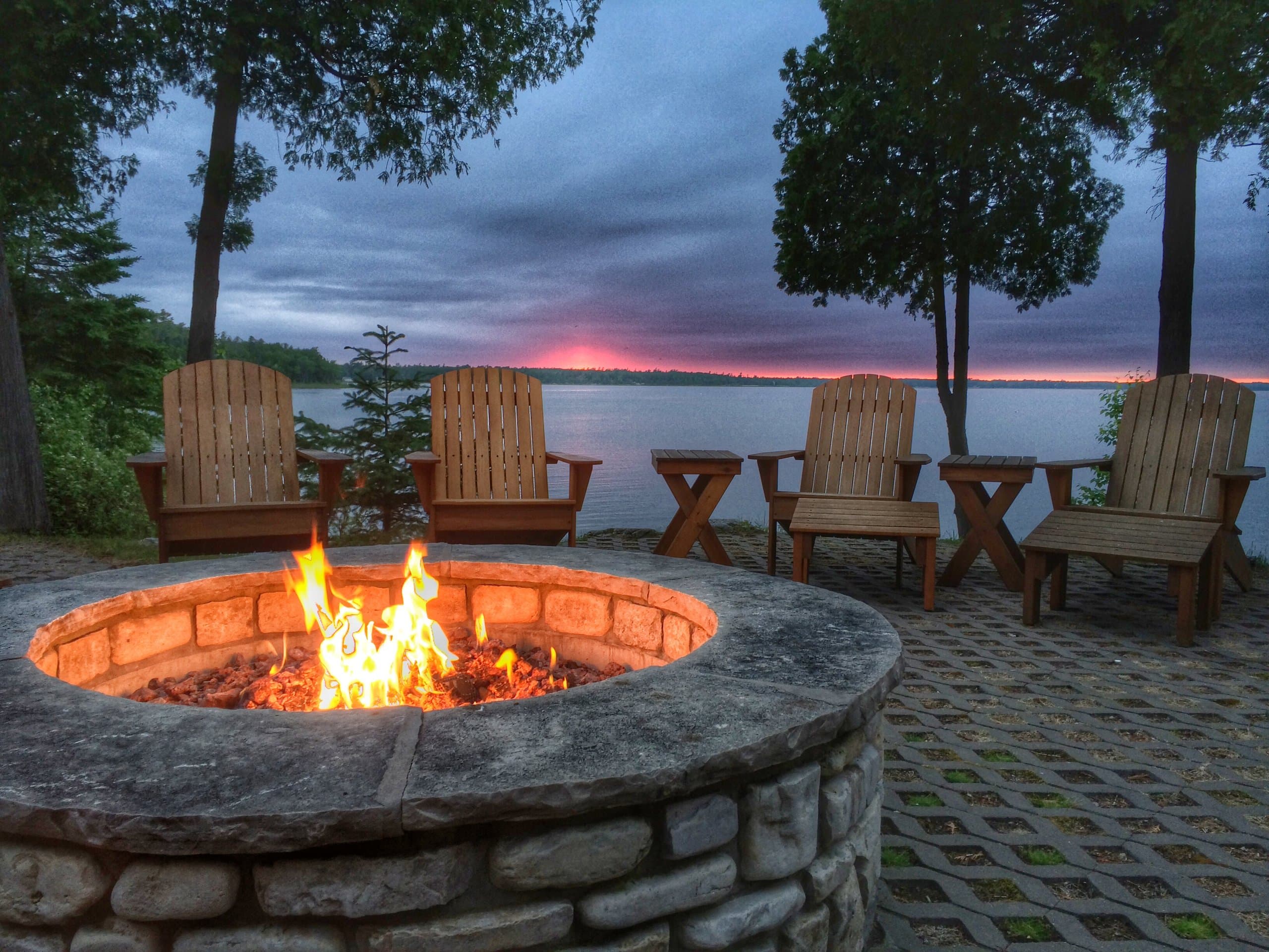 Stone fire pit and Adirondack chairs on a lakeside patio at sunset