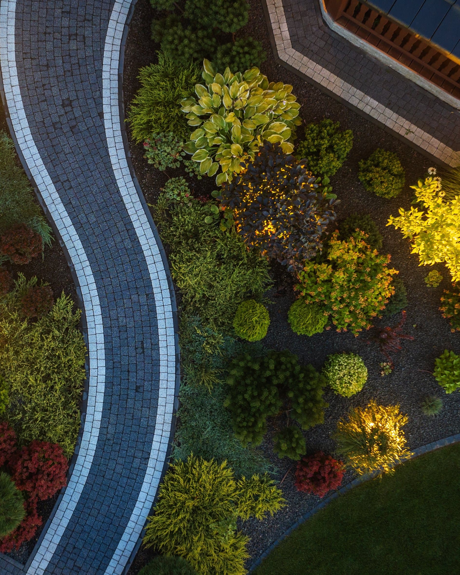 Aerial night view of a lit curved paver path winding through garden beds